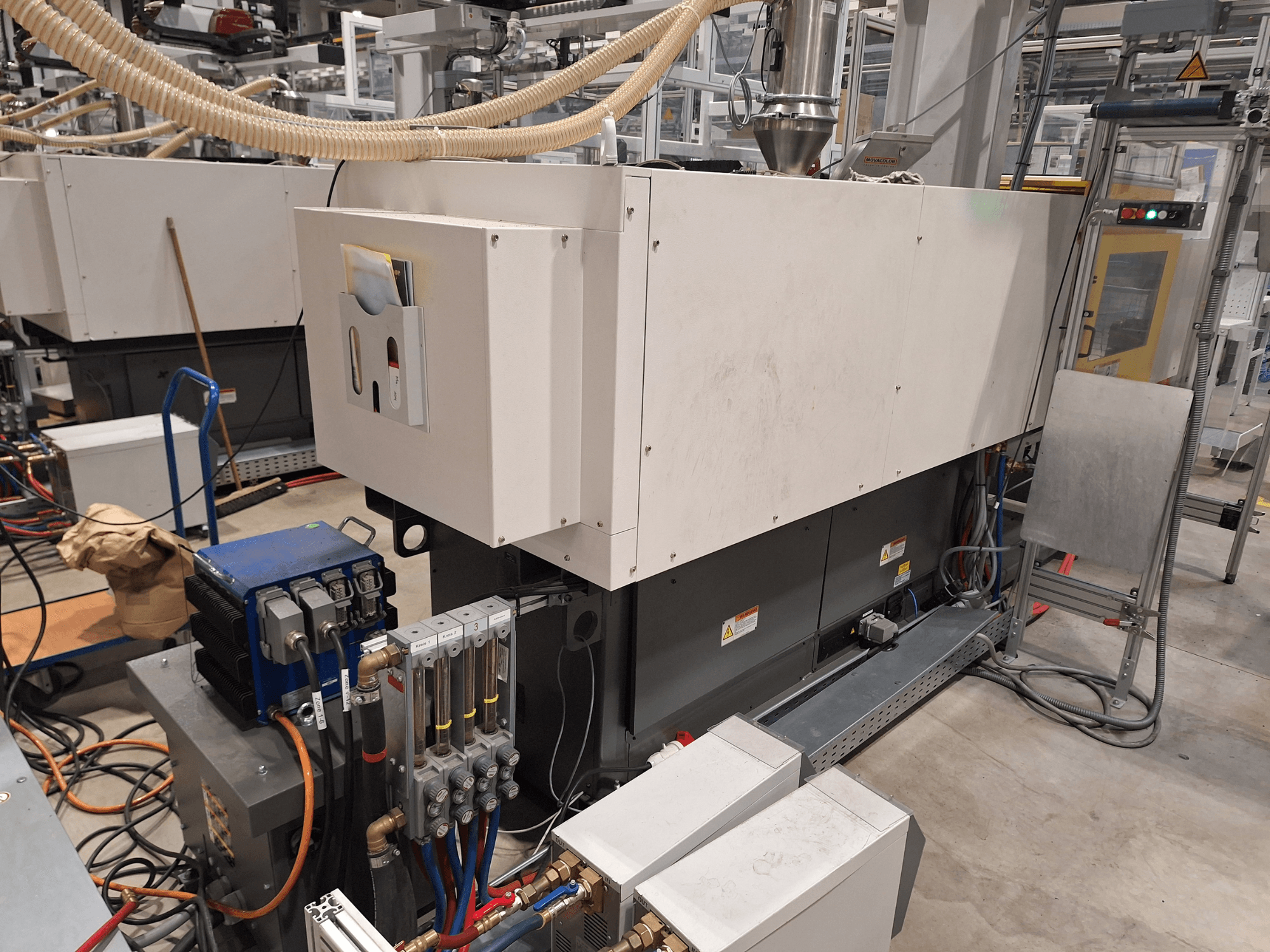 Industrielle Ausrüstung in einer Fabrik; umfasst eine große Maschine mit Bedienfeld, Schläuchen und elektrischen Anschlüssen, Seitenansicht.